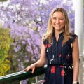 A young woman wearing a navy floral dress stands holding a railing with a flowering jacaranda tree in the background. She has blonde hair and is smiling.